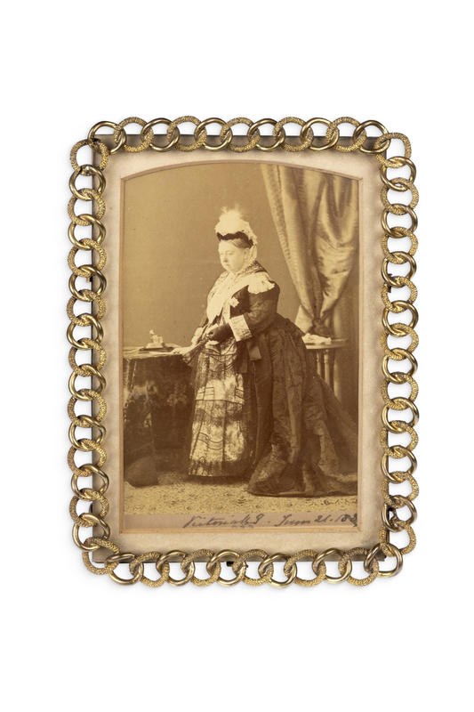 A sepia portrait of Queen Victoria standing next to a small table, in front of a curtain. The photograph is in a gold picture frame made of a chain.
